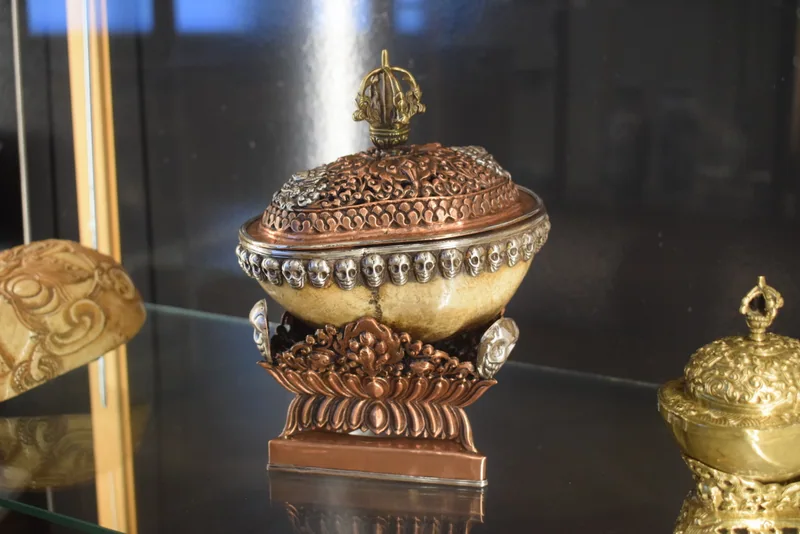 A mounted kapala with a pale rounded bowl, domed openwork lid and three-sided stand, photographed in a museum display.
